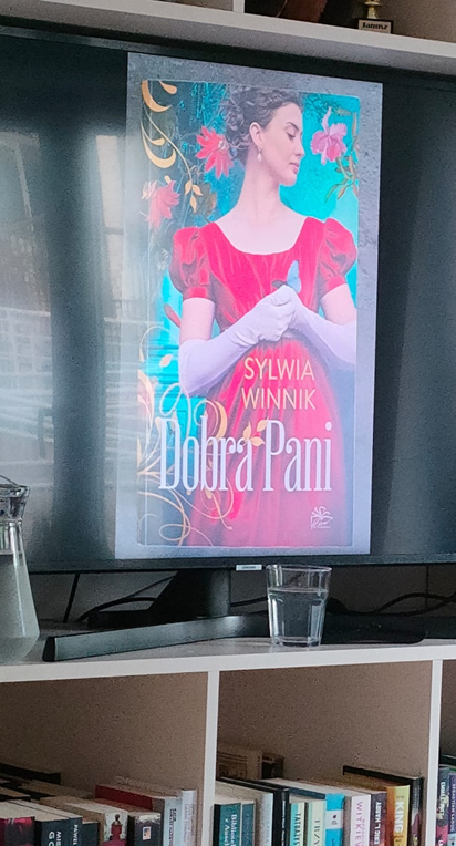 Zdjęcie_nr_13_przedstawia_wyświetlony_na_telewizorze_plakat_kobiety_w_czerwonej_sukience_otoczonej_roślinami_podpisany_Dobra_Pani.png
