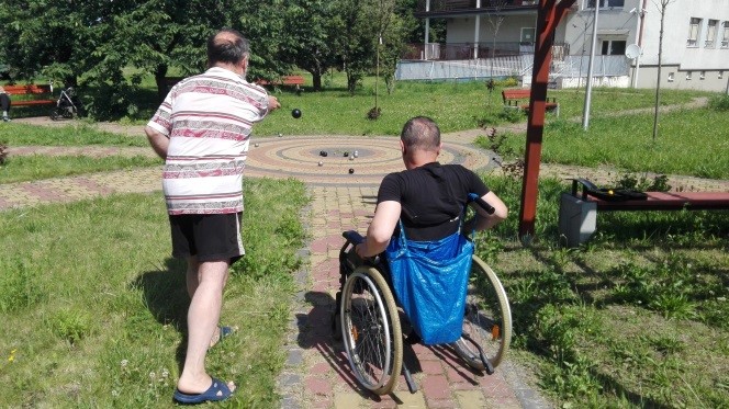 Zdjęcie przedstawia dwóch mieszkańców DPS Karmelkowa grających w ulubioną grę bule. Znajdują się w otoczeniu zieleni. Na drugim planie widać kawałek budynku DPS.