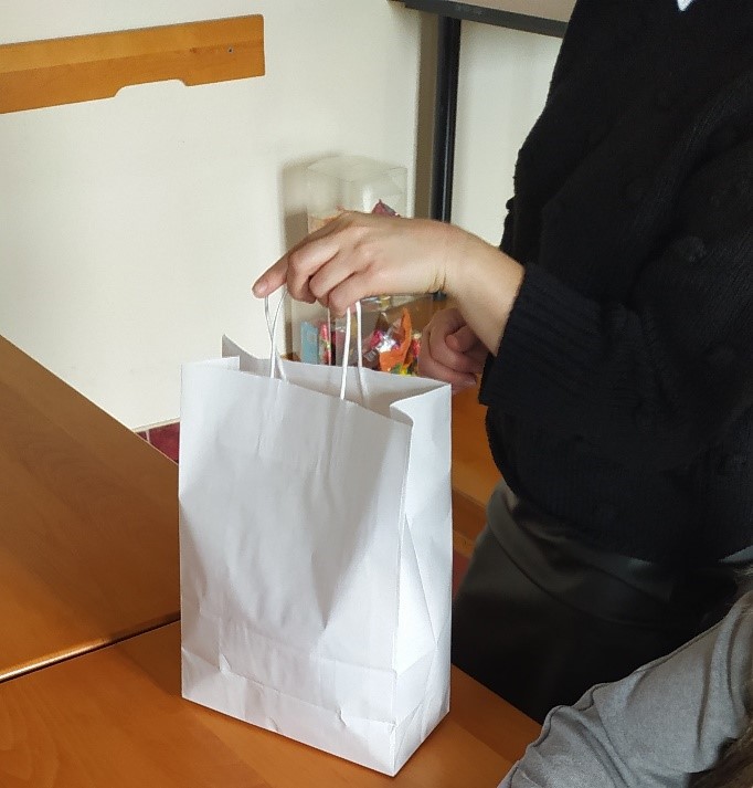 Na zdjęciu biała papierowa torba trzymana w dłoni kobiety, na tle brązowego stołu