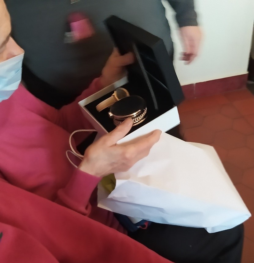 Na zdjęciu kobieta w różowym swetrze trzymająca pudełko z biżuterią i zegarkiem. Na kolanach leży biała, papierowa torba. W tle stojąca osoba