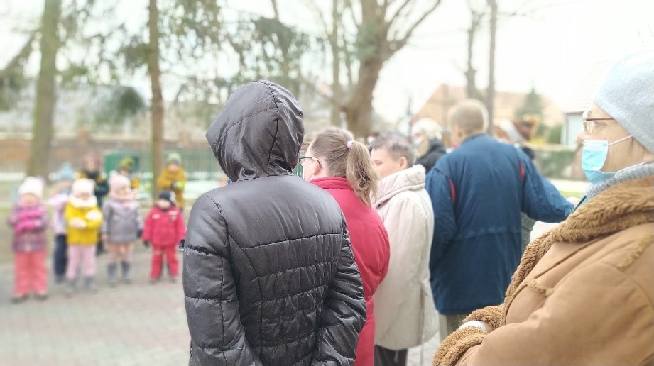 Na zdjęciu grupa ludzi w kurtkach, czapkach stojąca na dworze. W tle grupa dzieci i wysokie drzewa