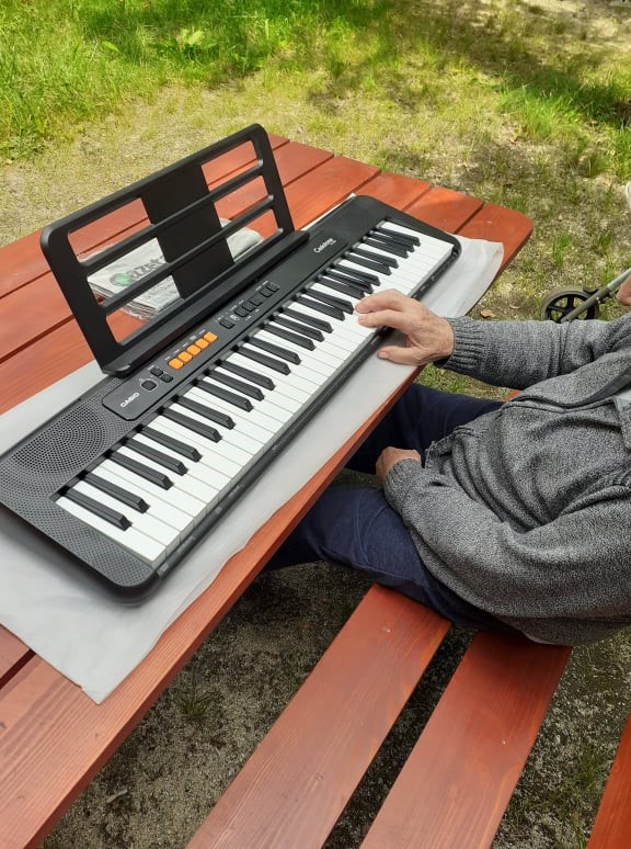 Fotografia przedstawia Pana siedzącego na brązowej, drewnianej ławce przy ogrodowym stole. Na stole jest ustawiony instrument muzyczny - keybord, na którym Klient gra prawą ręką