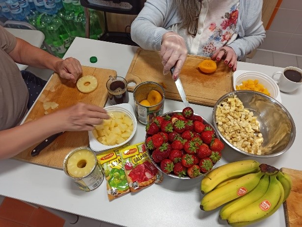 Na głównym planie mamy sezonowe owoce: banany, truskawki, brzoskwinie, ananasy. Mieszkańcy siedzą przy stole krojąc owoce na drewnianych deskach.