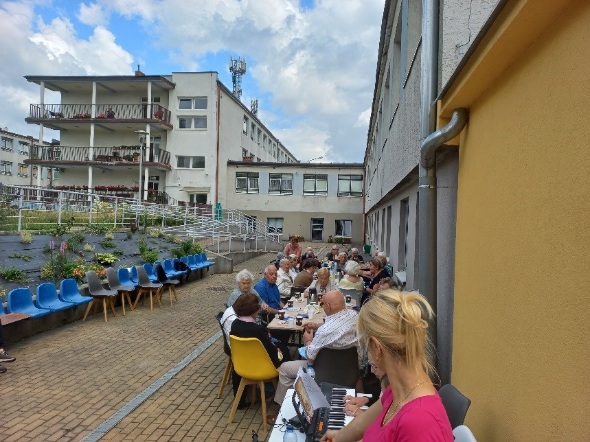 na fotografii widzimy na zewnątrz wspólnie siedzących Seniorów, przy stołach, rozmawiają