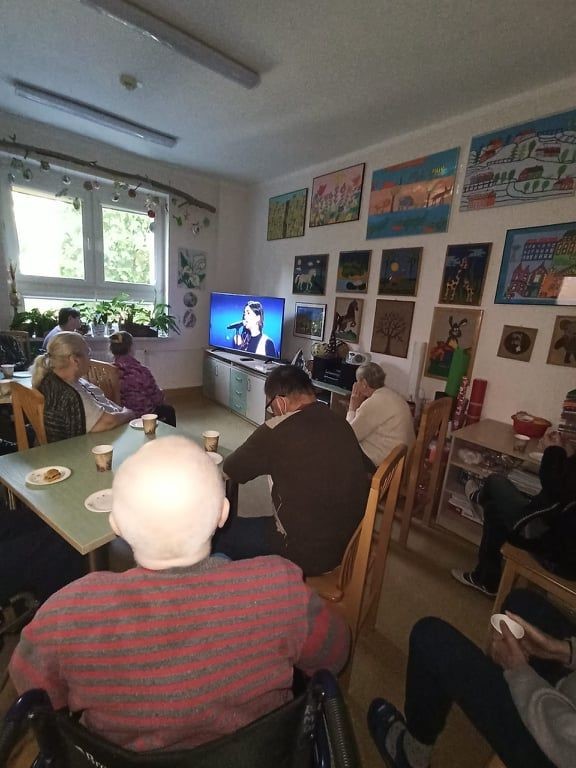 mieszkańcy siedzący przy stołach i oglądający występ na ekranie telewizora.