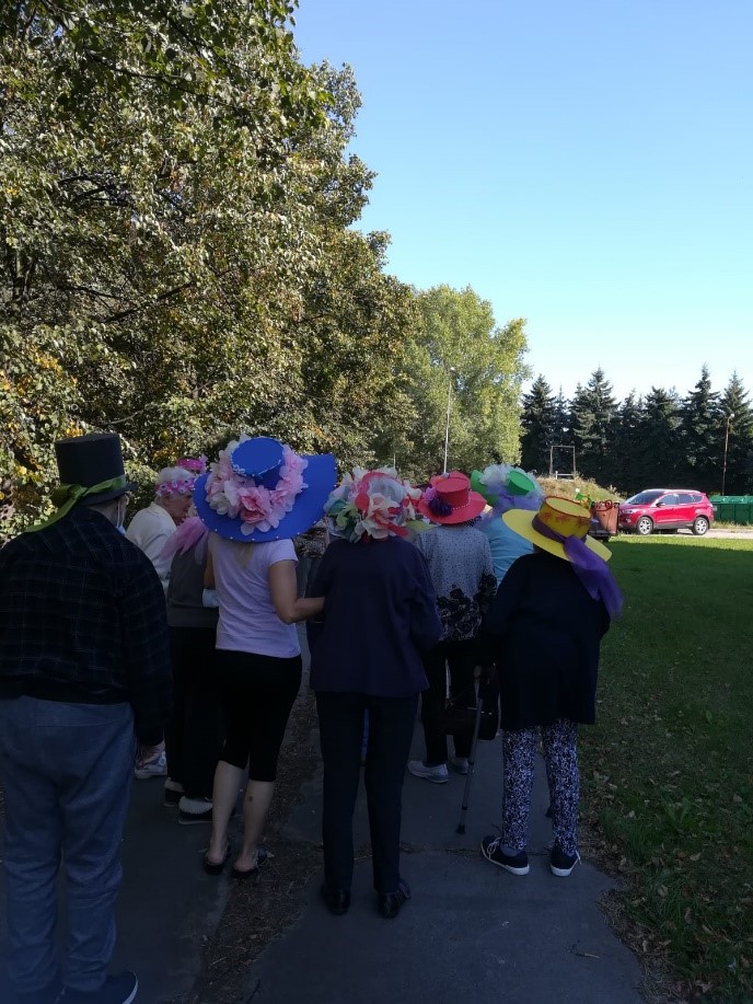 Teren zewnętrzny DDP. Klienci i klientki spacerują dookoła ośrodka w ramach „Marszu Kapeluszy”. Każdy nosi na głowie dekoracyjny, własnoręcznie ozdobiony kapelusz