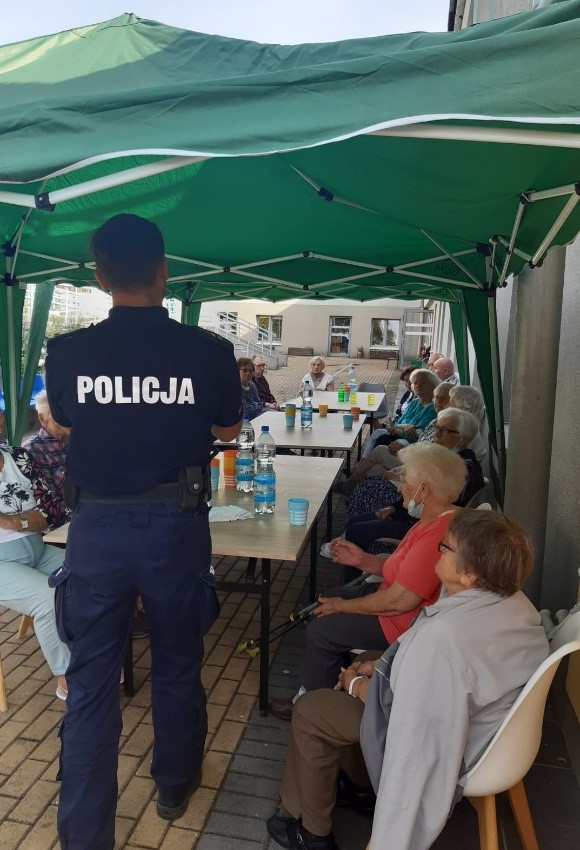 Teren zewnętrzny DDP. Na pierwszym planie stoi Funkcjonariusz Policji i przeprowadza z Klientami Ośrodka pogadankę dotyczącą bezpieczeństwa seniorów. Na dalszym planie Klienci DDP siedzą przy stołach, na których ustawione są napoje