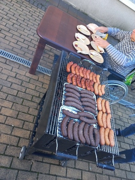 Przedstawia grilla na którym leży kiełbasa oraz kaszanka. Obok grilla jest rozłożony stolik a na nim tacki z chlebem, ketchupe i musztarda.