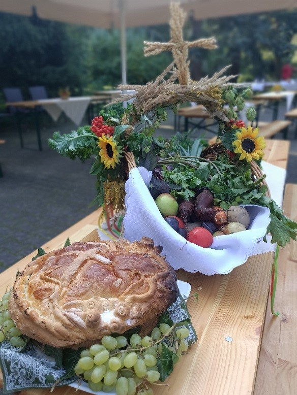 Na zdjęciu widać bochen chleba, kosz z warzywami i owocami ozdobiony polnymi kwiatami oraz wyplecionym z kłosów zboża krzyż.